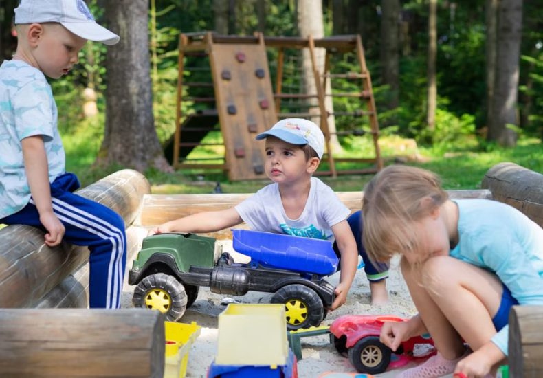 Three children engaged in imaginative play in an outdoor sandbox with toys.