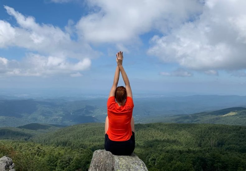 A woman in a red shirt relaxes on a mountain peak with arms raised, enjoying the scenic view.